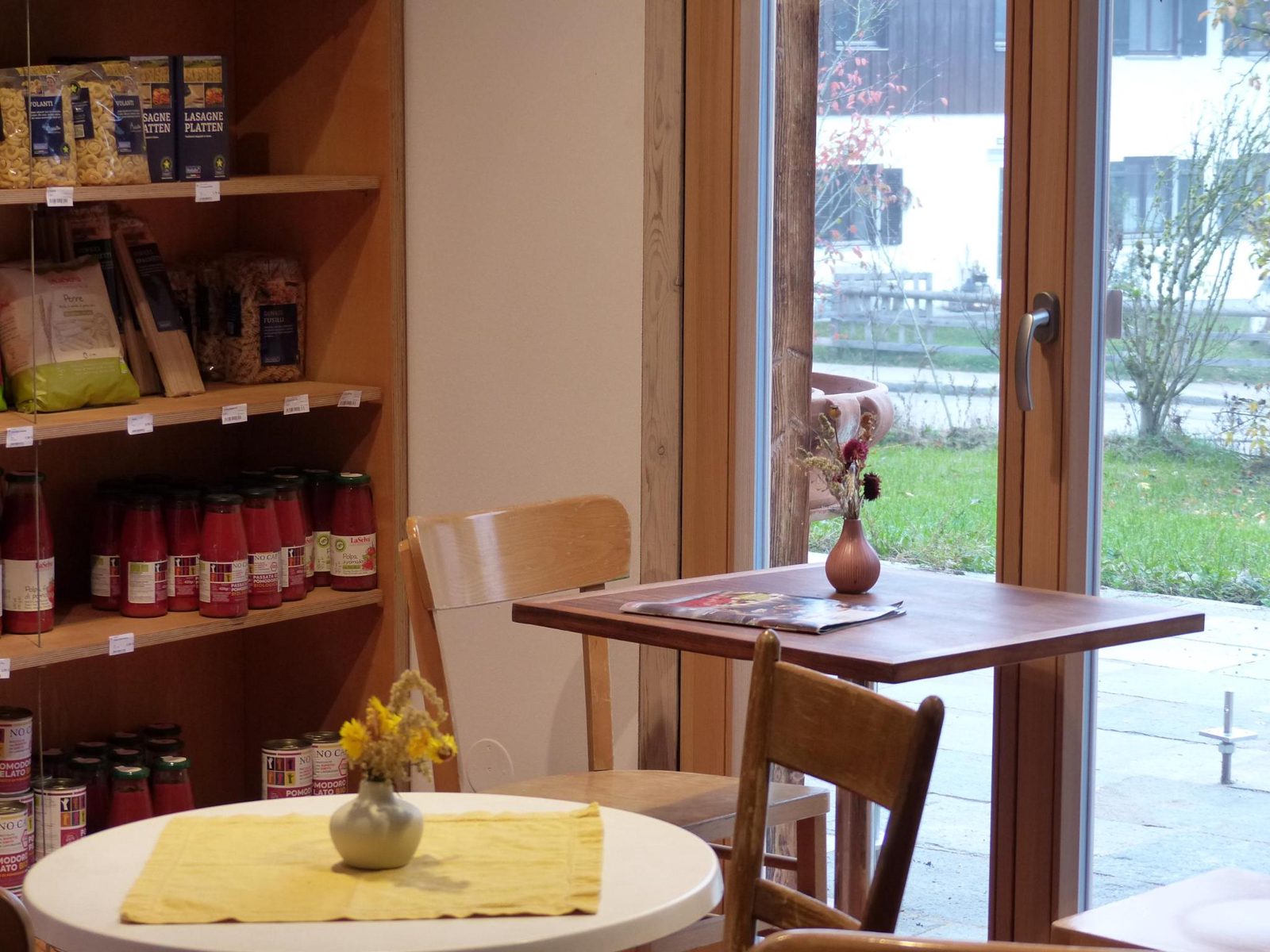 Café-Tisch am Fenster mit Trockenblumen, daneben Regale mit Pasta und Tomaten